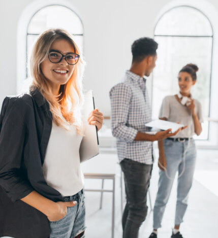 Elegant business-lady in trendy black jacket holding laptop and smiling. Portrait of cheerful blonde secretary and tall african office worker on background.